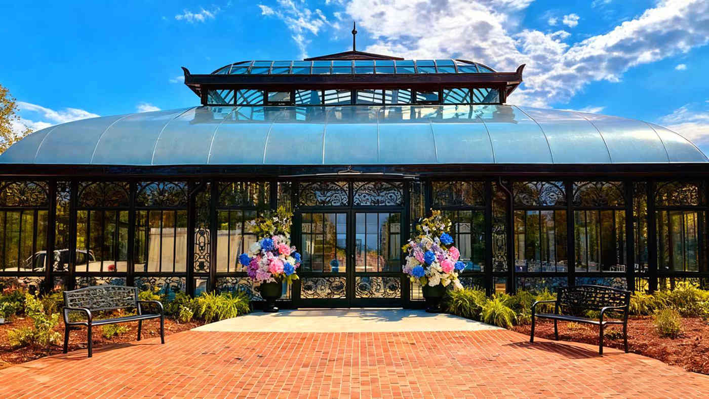 Victorian conservatory wedding venue, black ornate metal frame sunroom with curved glass roof, brick walkway and floral decorations for outdoor event space